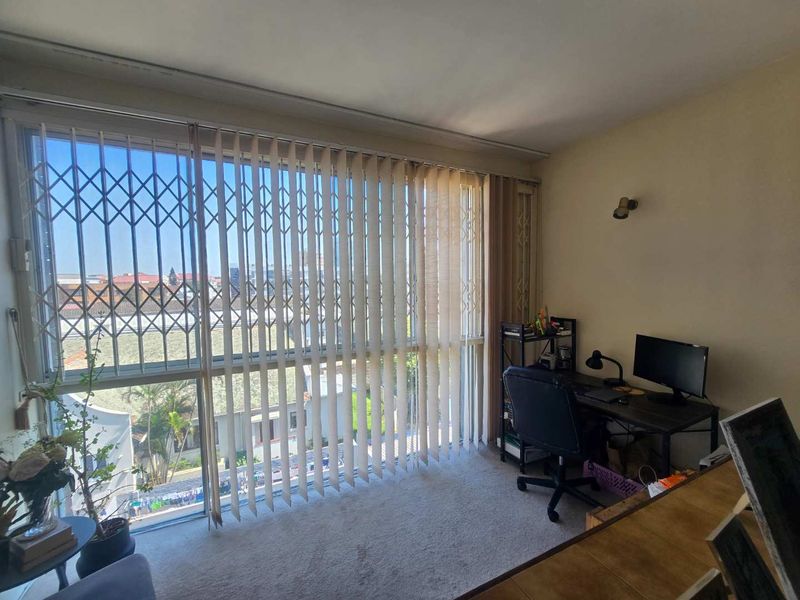 A bedroom featuring carpet flooring, vertical blinds, and a security gate over the window, offering natural light and a view of the surrounding area.