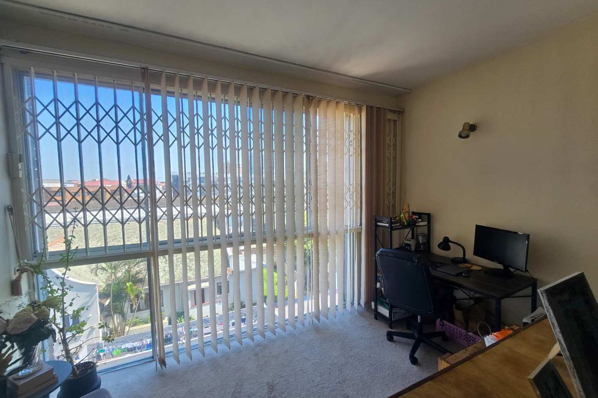 A bedroom featuring carpet flooring, vertical blinds, and a security gate over the window, offering natural light and a view of the surrounding area.