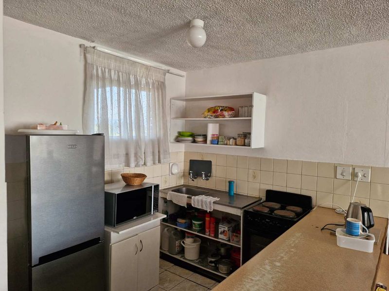 A compact kitchen featuring a stainless steel sink, tiled backsplash, electric stove, and open shelving, requiring modernization of finishes and cabinetry.