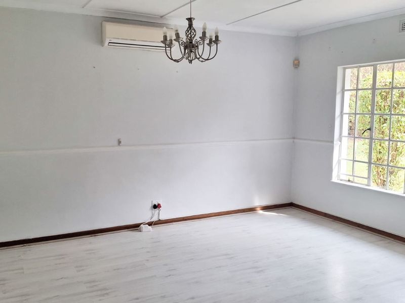 A bright, empty living room featuring light-toned laminate flooring, a wall-mounted air conditioning unit, and a classic chandelier.