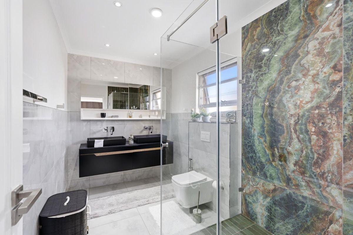 Modern bathroom featuring a double vanity, wall-mounted toilet, glass shower enclosure, and a striking green stone feature wall.