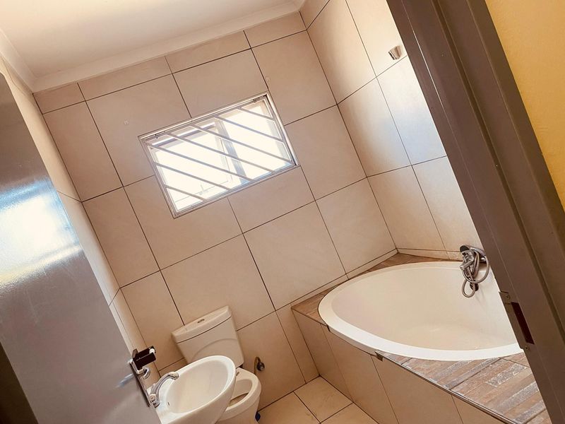 A clean, tiled bathroom featuring a built-in bathtub, pedestal basin, and toilet, with a window fitted with security bars.