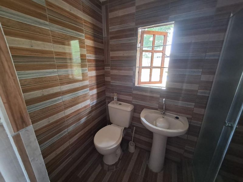 A functional bathroom featuring full-height wood-look wall tiling, a pedestal sink, a standard toilet, and a shower enclosure with a small casement window.