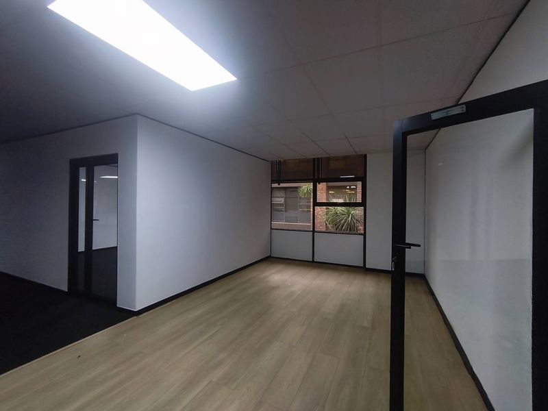A bright, vacant office space featuring modern laminate flooring, glass partition doors, and a suspended ceiling with recessed lighting.