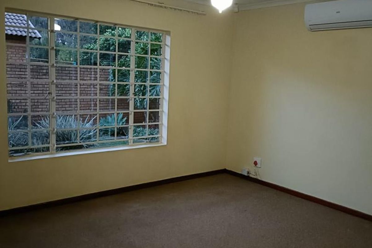A spacious, empty bedroom featuring neutral walls, a ceiling fan, and a wall-mounted air conditioning unit, overlooking the garden through a large window.