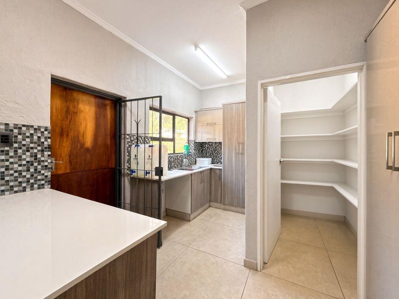 A functional scullery area featuring built-in cabinetry, a tiled backsplash, a stainless steel sink, and an adjacent walk-in pantry with ample shelving.