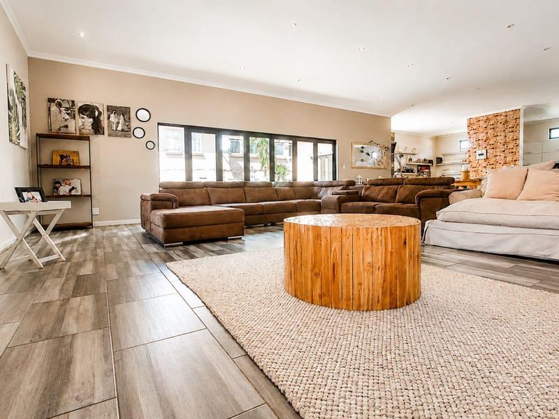 Spacious open-plan living area featuring wood-look tile flooring, recessed lighting, and bi-fold doors leading to the outdoors, with an adjacent kitchen and brick feature wall.