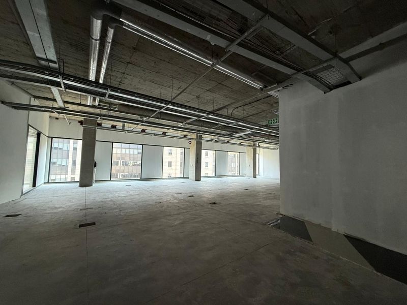 Spacious open-plan office area featuring an industrial aesthetic with exposed concrete ceilings, ducting, and large windows providing ample natural light.