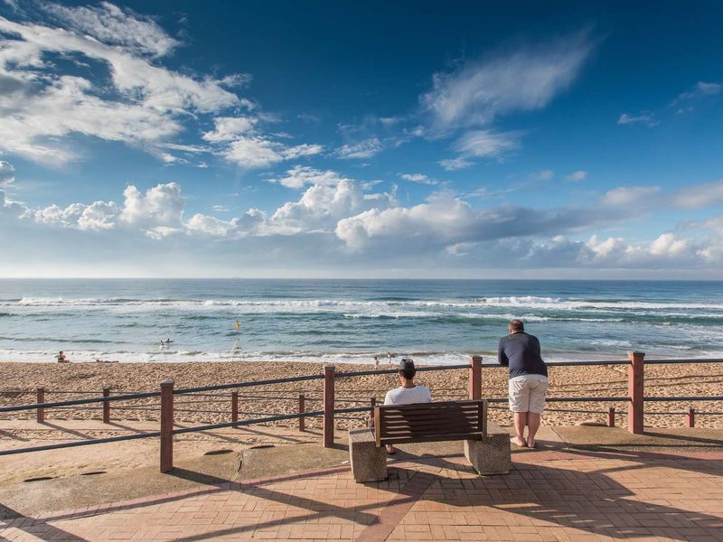 A scenic view of the ocean and beach from a paved promenade featuring a protective railing.