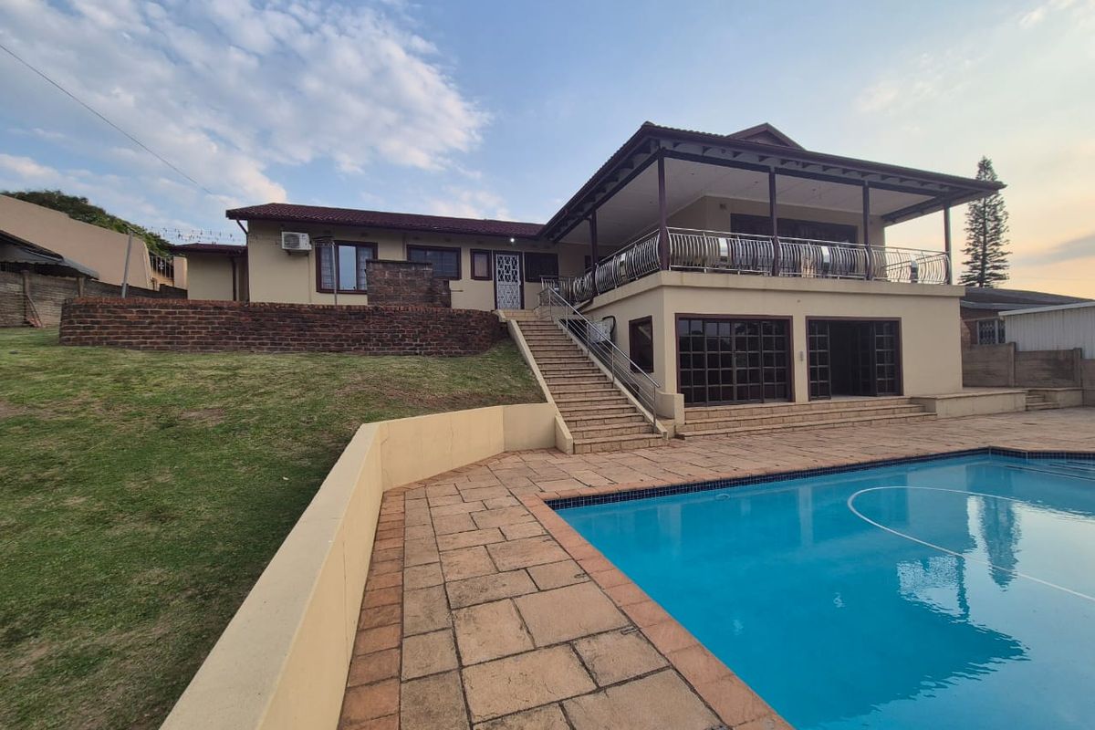 Rear exterior view featuring a swimming pool, paved deck, covered patio, and an elevated balcony with stairs leading to the garden area.