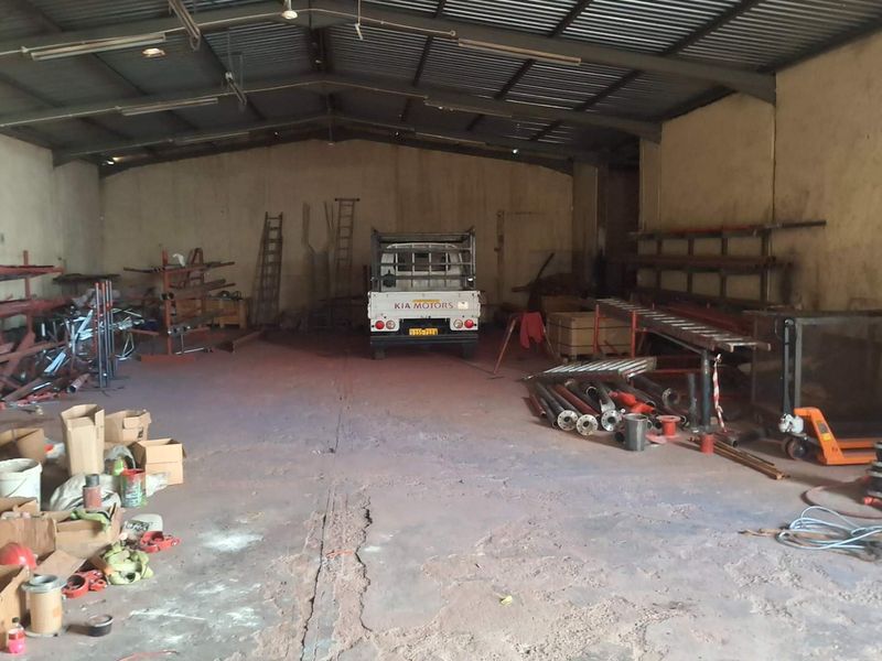 Spacious industrial utility area featuring high ceilings with exposed steel trusses, concrete flooring, and fluorescent lighting, suitable for storage or workshop operations.
