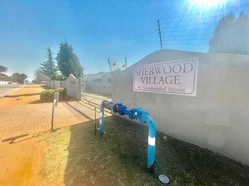 The entrance to Sherwood Village, featuring a boundary wall with electric fencing, a metal gate, and visible external water infrastructure.