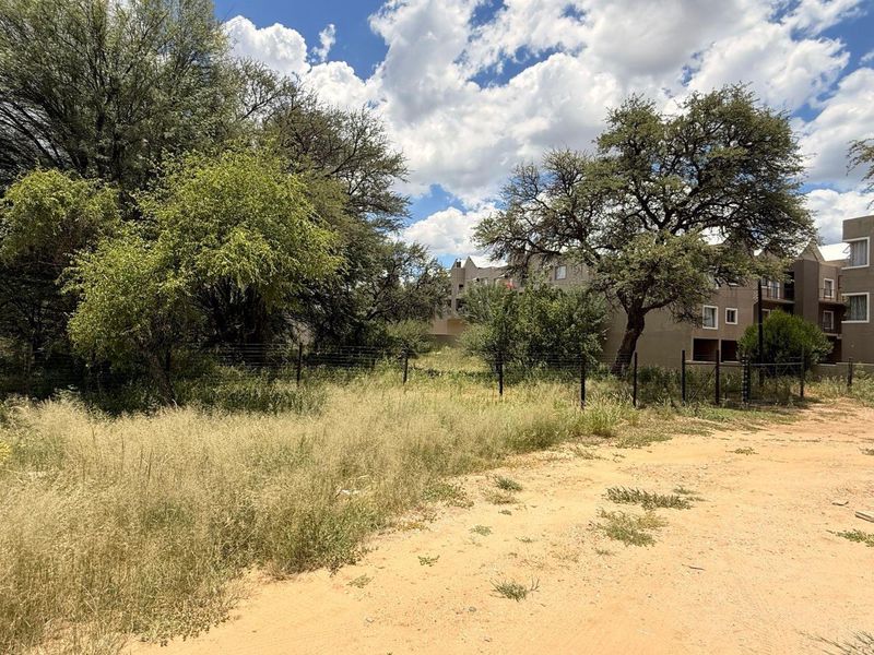 A view of a vacant residential plot featuring natural vegetation, mature trees, and existing wire mesh perimeter fencing.
