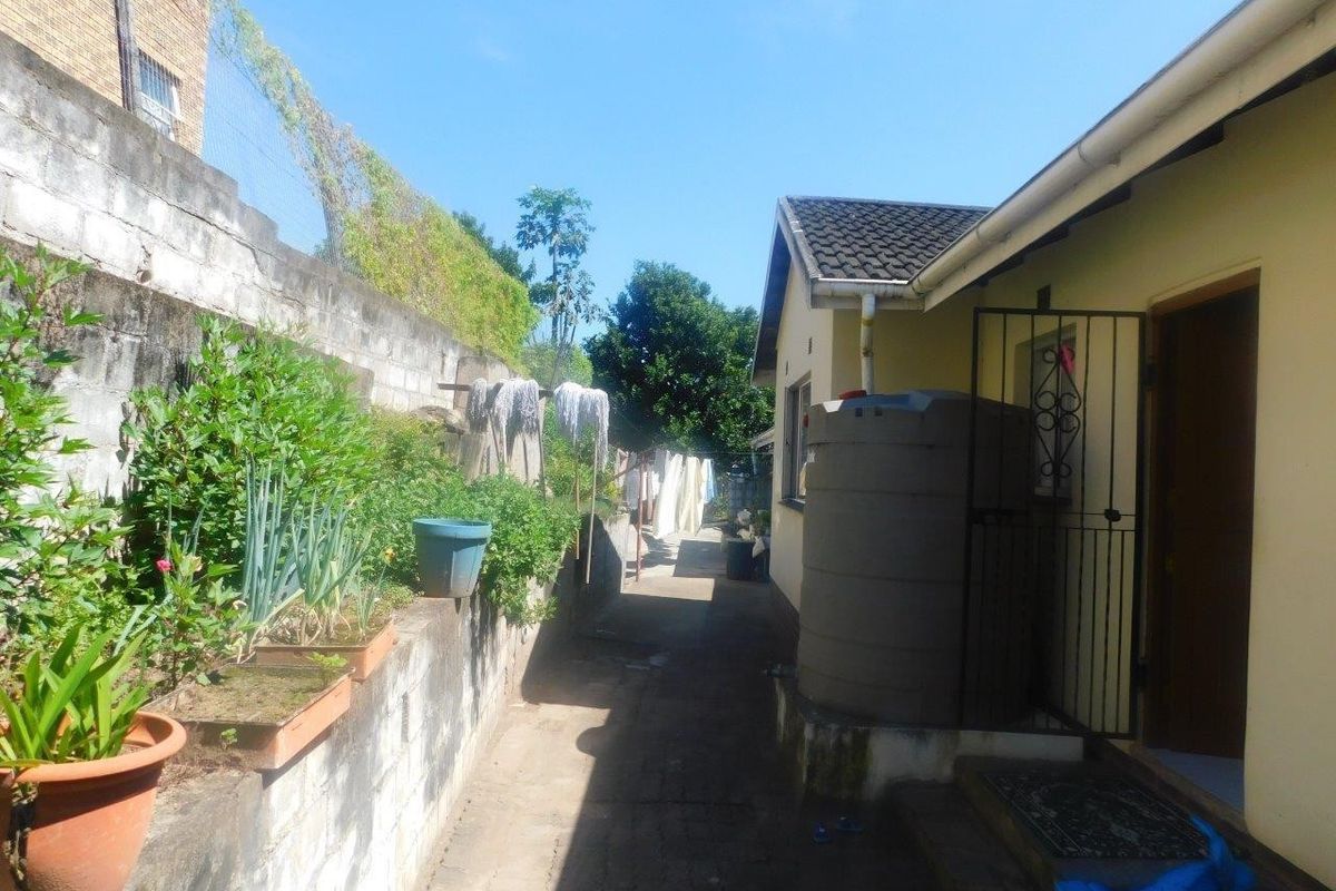 A narrow rear exterior pathway featuring a concrete boundary wall, a water storage tank, and a security gate entrance to the home.