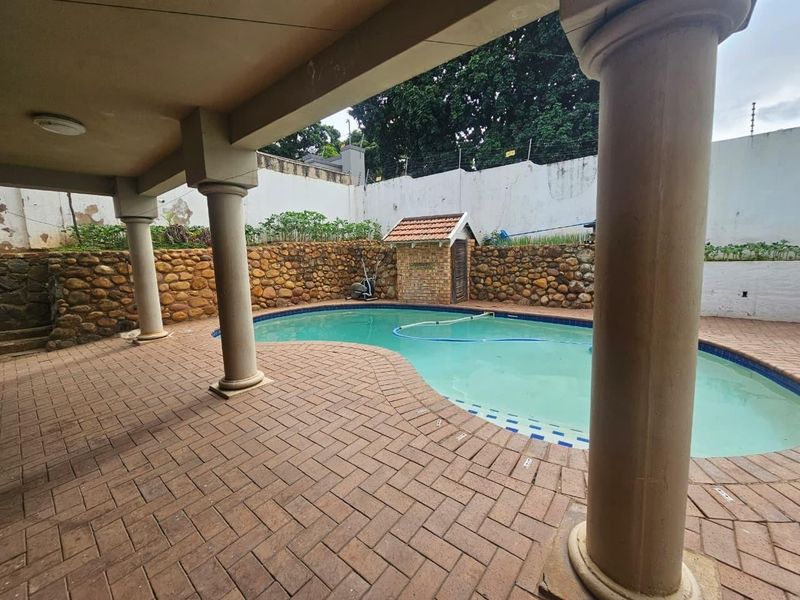 Outdoor pool area featuring a covered patio with concrete columns, brick paving, and a stone retaining wall, enclosed by a perimeter wall with electric fencing.