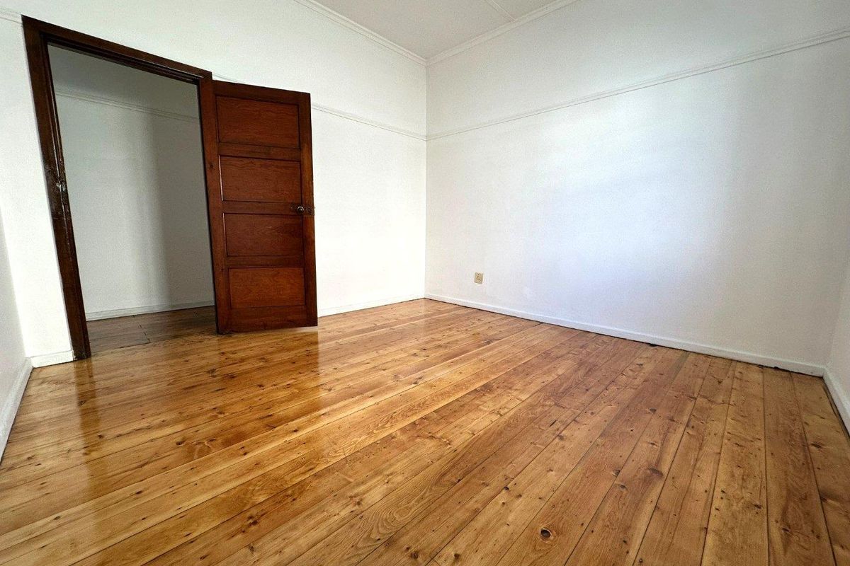 A bright, empty bedroom featuring polished timber flooring, white walls with picture rails, and a classic wooden door leading to an adjacent room.