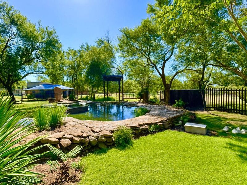 A natural-style swimming pool area featuring stone masonry edging, surrounded by a lush garden and mature trees on a farm property.