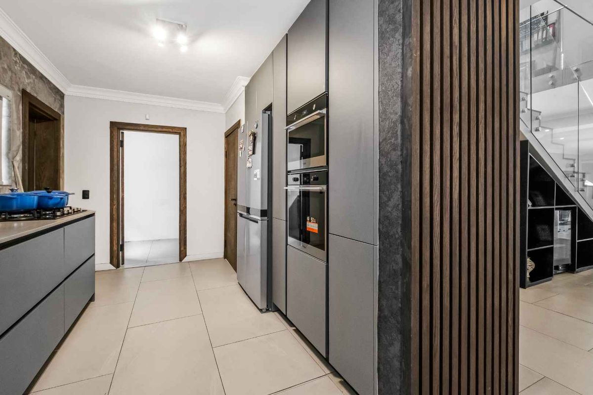Modern kitchen featuring sleek grey cabinetry, integrated appliances, and stylish timber slat wall paneling, leading to an open staircase with glass balustrades.
