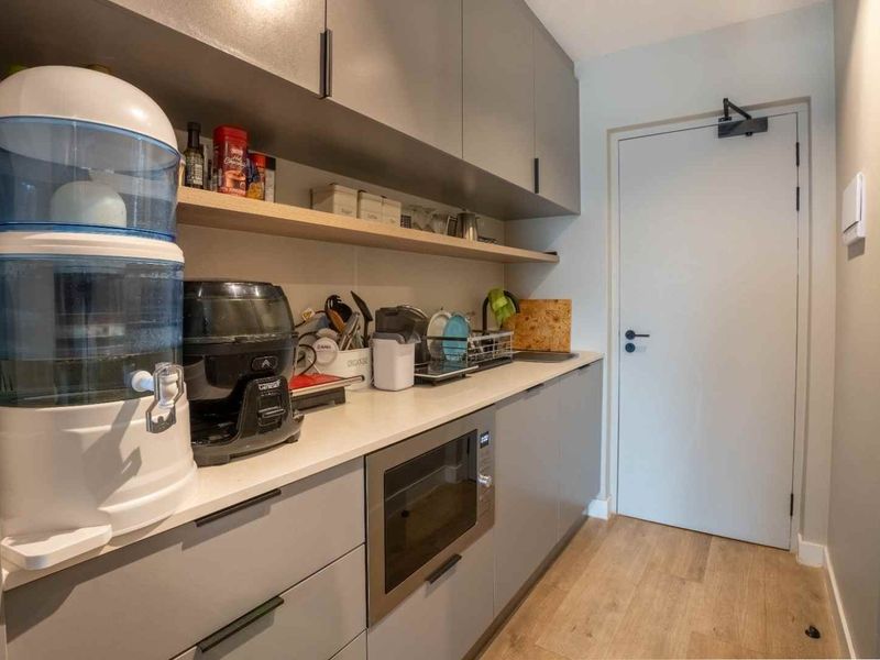 A modern, compact scullery featuring sleek grey cabinetry, stone countertops, built-in microwave, and open shelving, providing efficient additional storage and prep space.