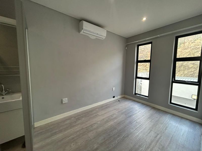 A bright, empty bedroom featuring modern laminate flooring, an integrated air conditioning unit, and an en-suite vanity basin area with large windows.