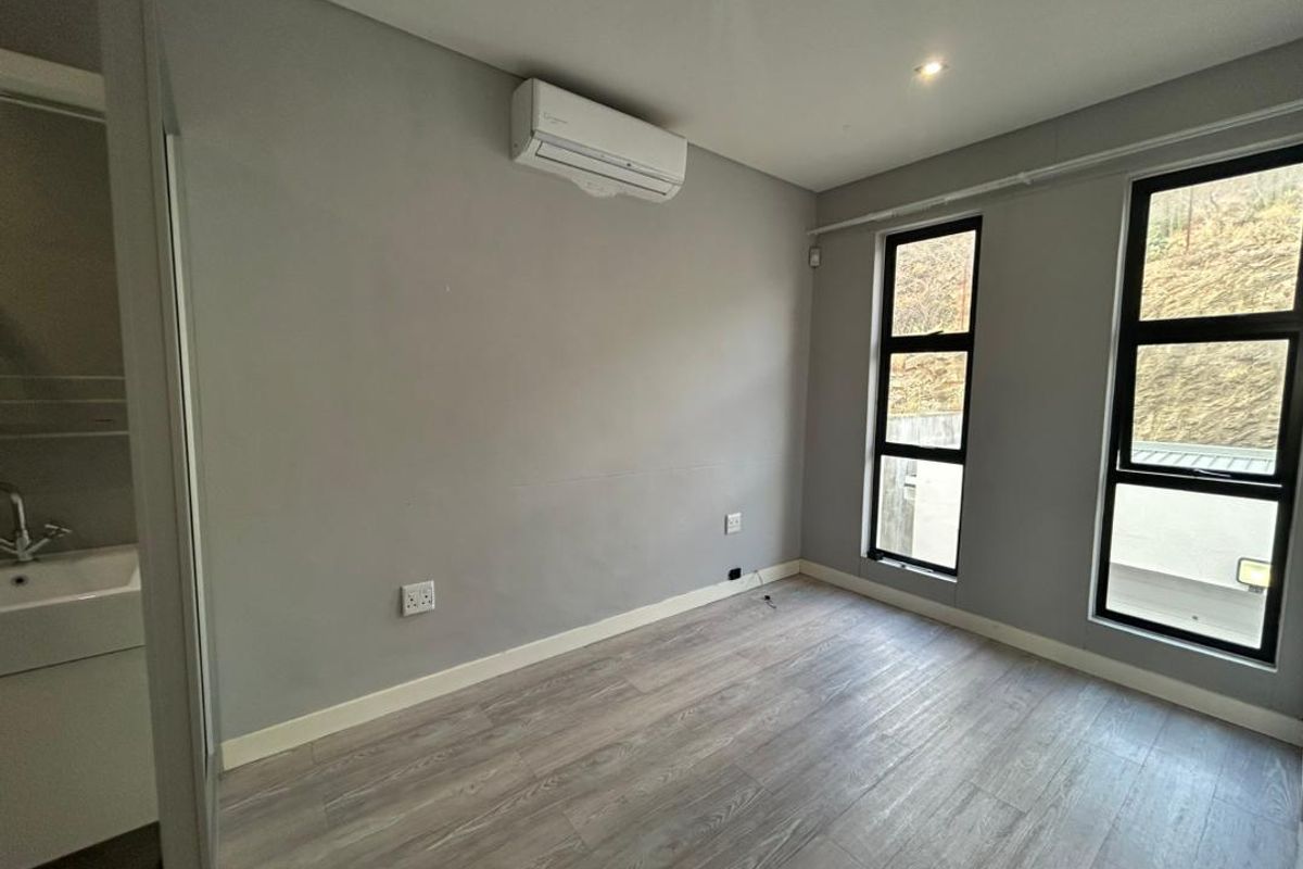 A bright, empty bedroom featuring modern laminate flooring, an integrated air conditioning unit, and an en-suite vanity basin area with large windows.