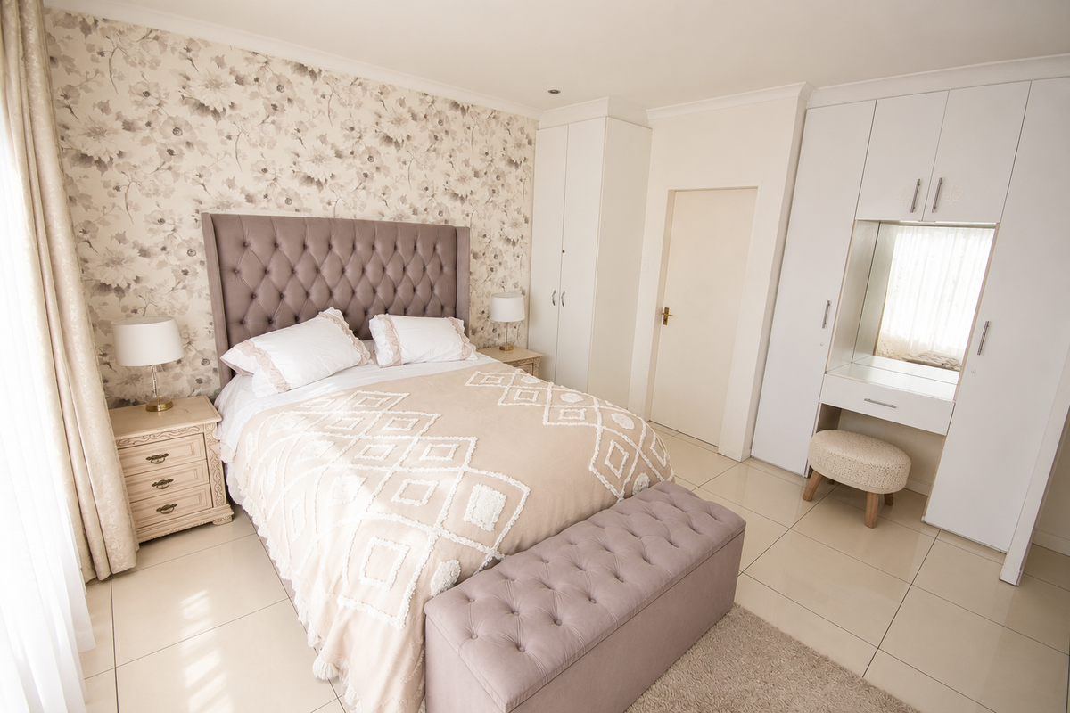 A well-maintained bedroom featuring built-in cupboards, a integrated vanity station, tiled flooring, and decorative wallpaper behind the bed.