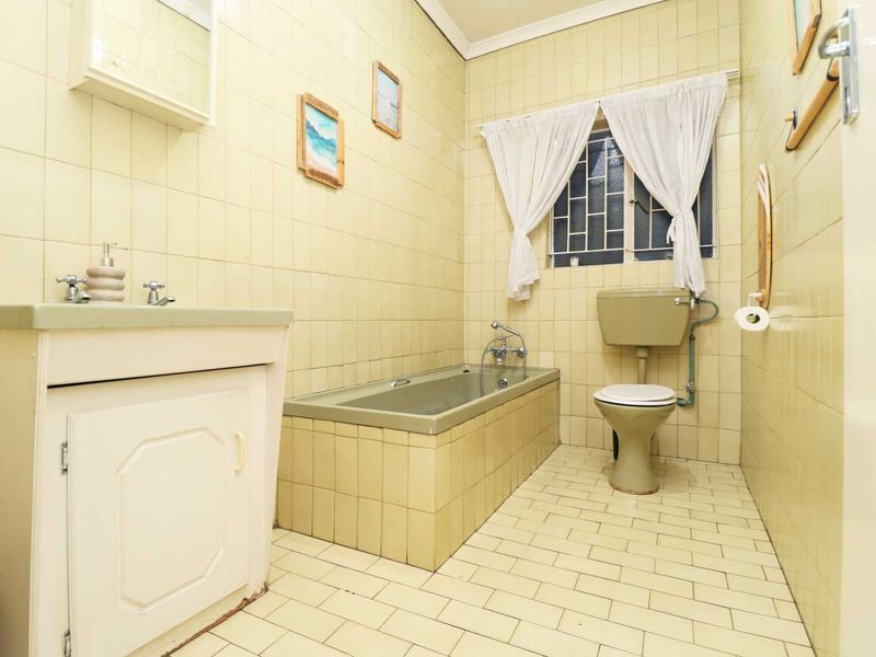 A dated bathroom featuring tiled walls and flooring, a built-in vanity, a bathtub, and a standard toilet with security bars on the window.