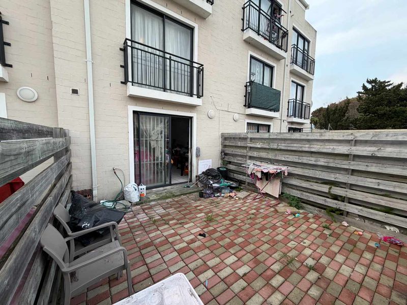 Rear exterior view of an apartment building featuring a paved patio area enclosed by timber fencing and access via sliding glass doors.