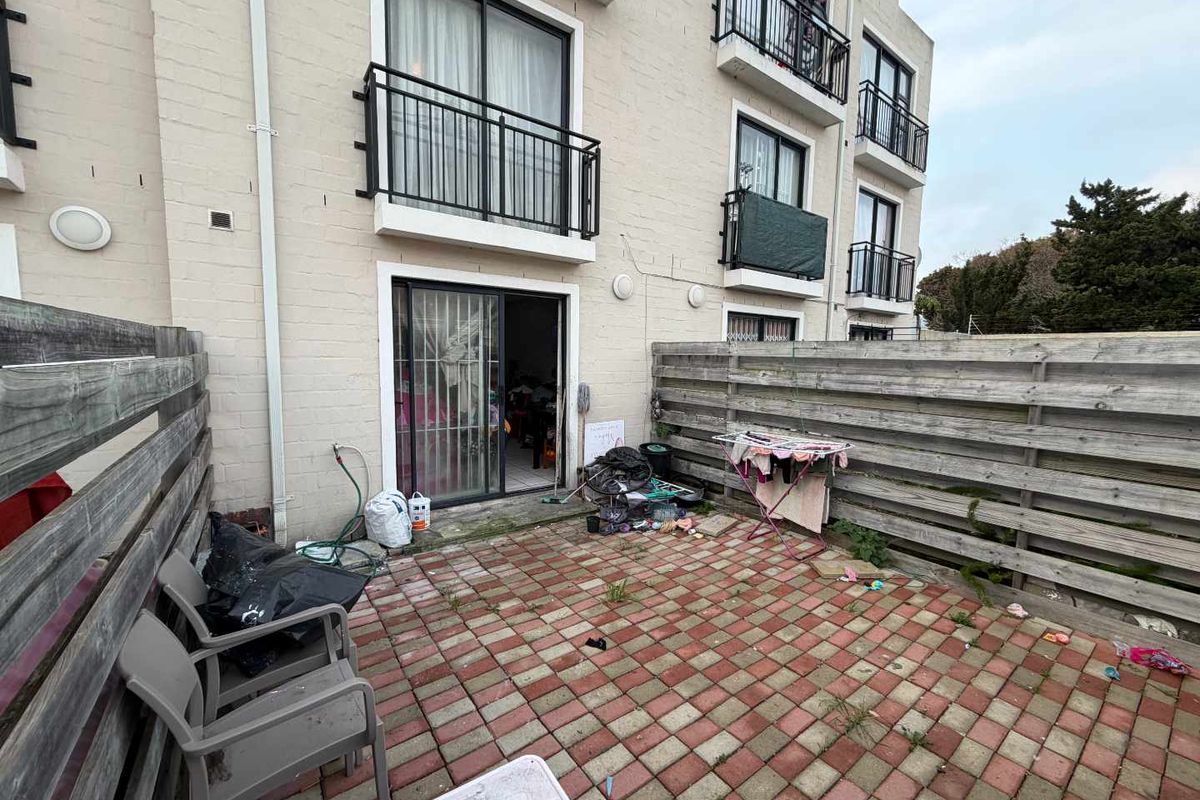 Rear exterior view of an apartment building featuring a paved patio area enclosed by timber fencing and access via sliding glass doors.