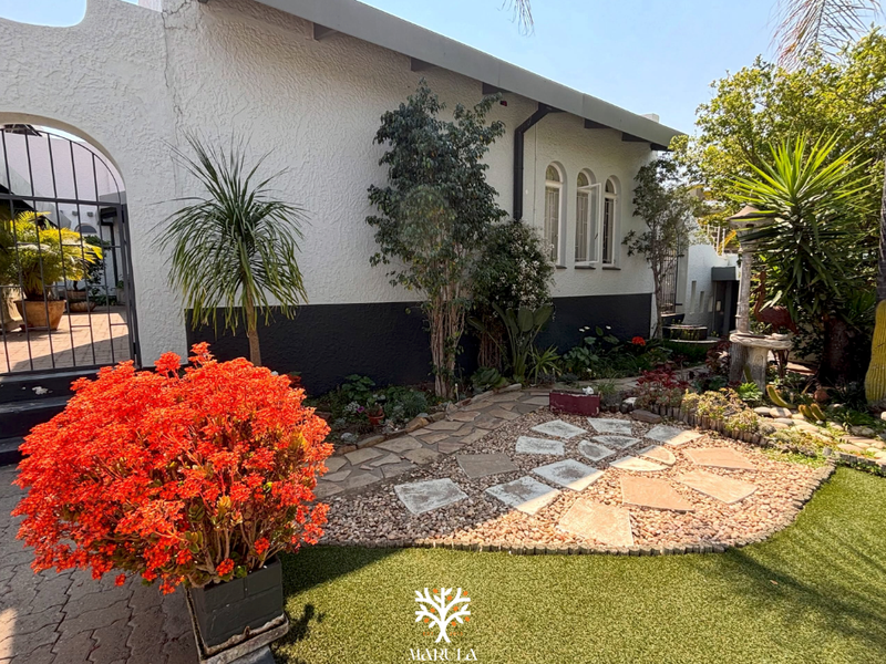 A well-maintained rear garden area featuring a stone-paved pathway, artificial lawn, and a charming arched metal gate leading to the side of the house.