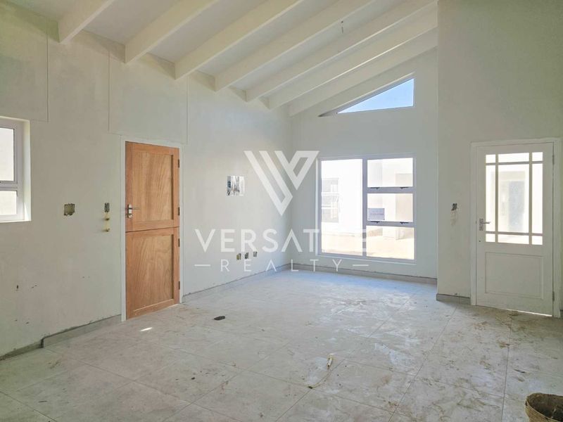 An empty, unfinished living room featuring high exposed beam ceilings, tiled flooring, and natural light from large aluminium windows.