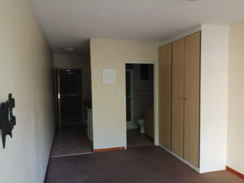 An empty living area featuring built-in cupboards, carpeted flooring, and direct access to a kitchen and bathroom area.