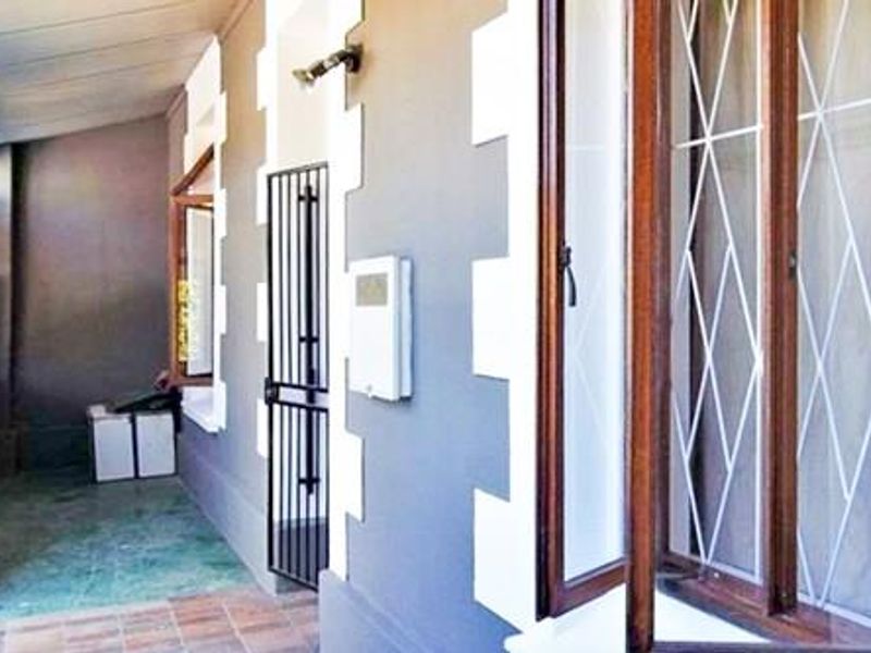 A covered patio area featuring tiled flooring, a security gate, and wooden window frames, providing a functional outdoor transition space.