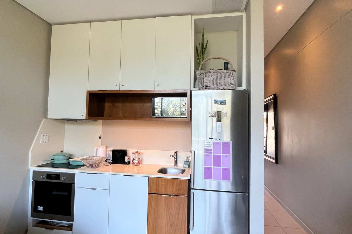 Compact kitchen featuring modern white and wood-tone cabinetry, integrated oven, cooktop, and stainless steel refrigerator, with a clean tiled floor and recessed lighting.