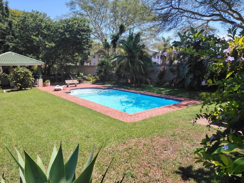 A well-maintained outdoor pool area featuring a rectangular swimming pool with brick coping, surrounded by a lush garden and a covered gazebo.