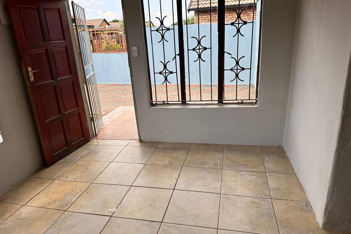 An empty interior room featuring tiled flooring, a wooden entrance door, and large windows fitted with security bars, providing direct access to the exterior.