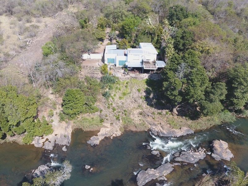 Aerial view of a farm property situated on a rocky riverbank, featuring a multi-level structure with metal roofing and an elevated deck.