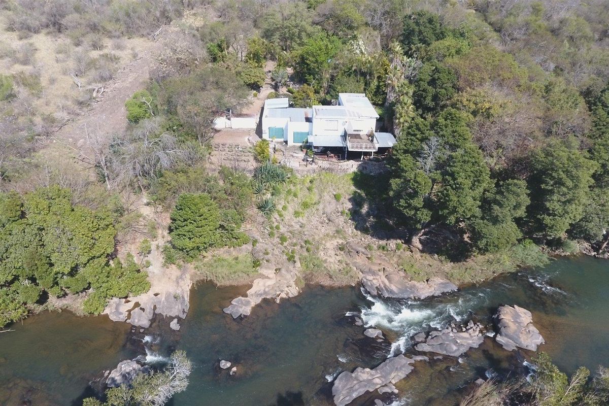 Aerial view of a farm property situated on a rocky riverbank, featuring a multi-level structure with metal roofing and an elevated deck.