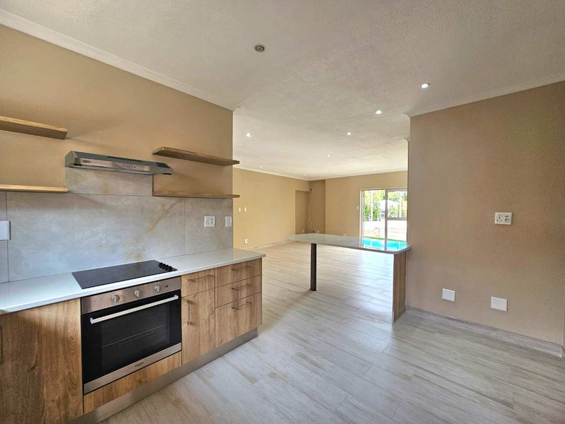 Modern open-plan kitchen and living area featuring wood-look cabinetry, built-in appliances, a breakfast bar, and neutral tiled flooring with direct access to the pool area.