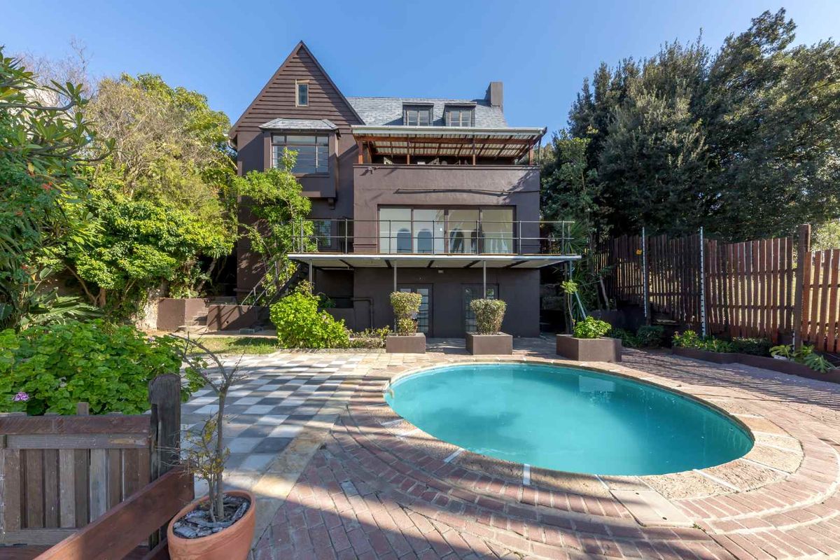 Rear view of a multi-story house featuring a swimming pool, paved patio area, and a balcony overlooking the garden.