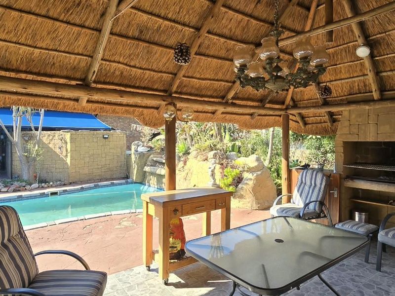 Covered thatched patio area featuring a built-in braai, overlooking a swimming pool and landscaped garden, ideal for outdoor entertaining.