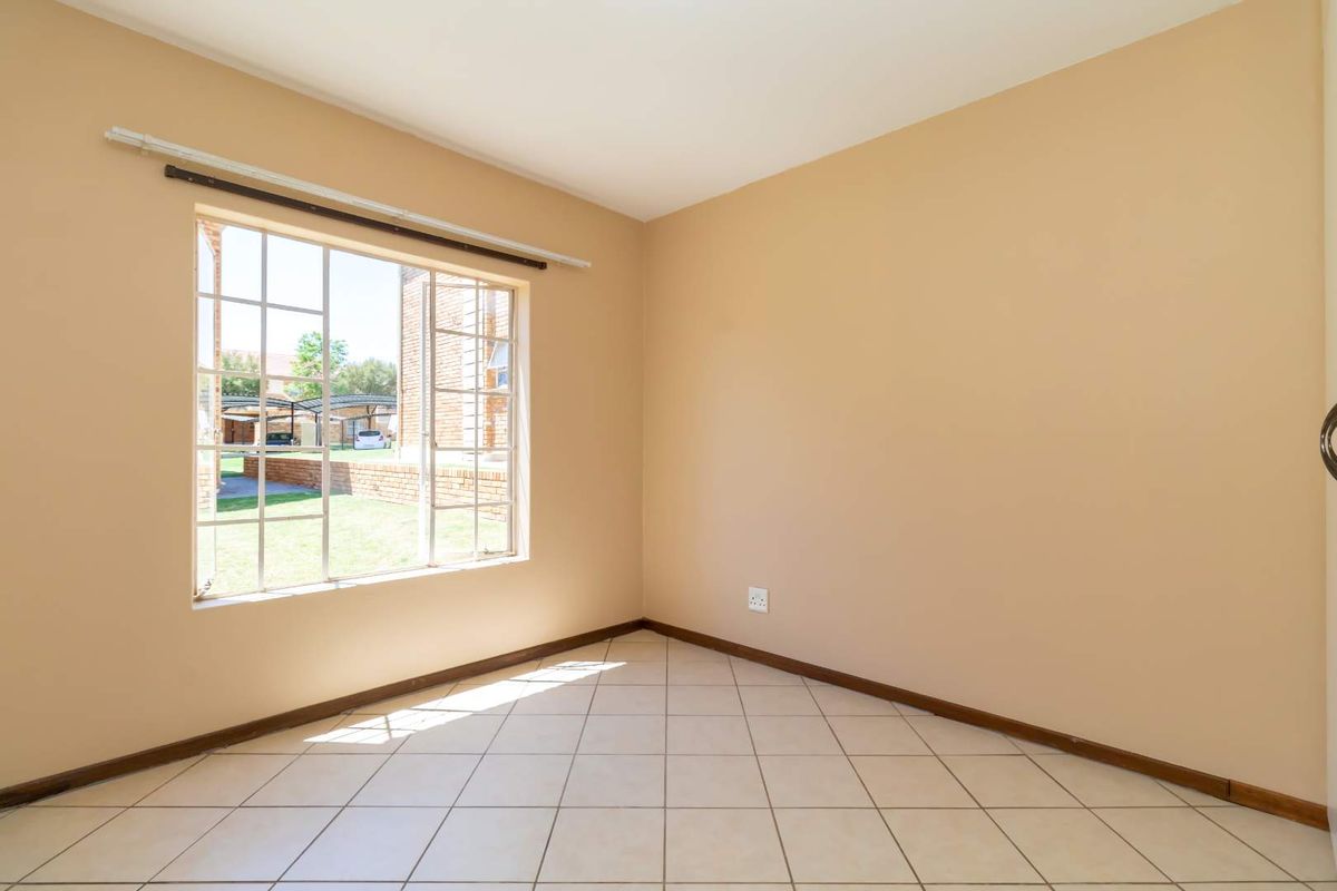 A bright, empty bedroom featuring neutral wall paint, tiled flooring, and a large window with security bars overlooking the complex grounds.