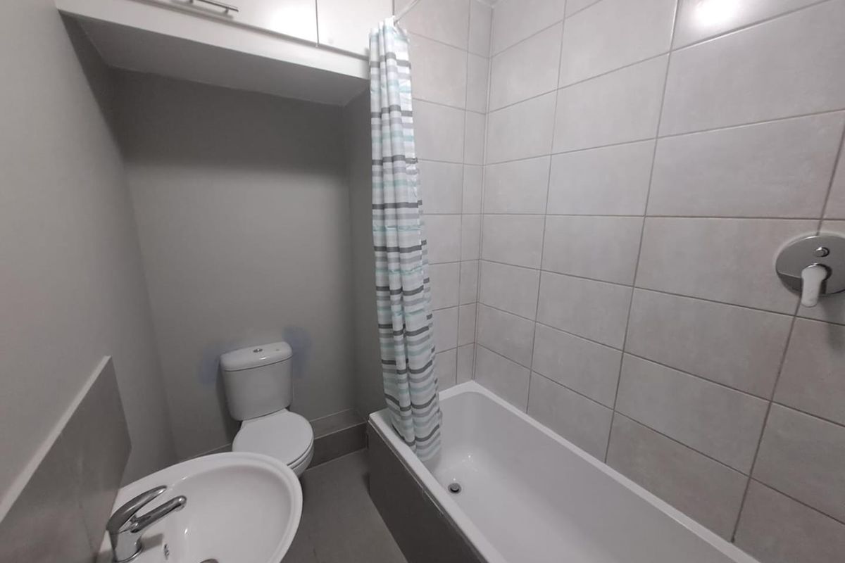 Compact bathroom featuring a white bathtub with shower head, pedestal basin, and toilet, finished with neutral wall tiling and overhead storage cabinetry.