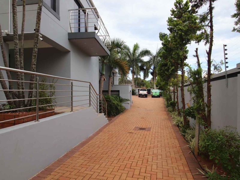 A paved driveway leading to the front of the property, featuring modern grey exterior walls, stainless steel balustrades, and secure perimeter fencing.