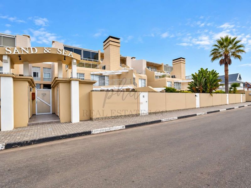 Front exterior view of the Sand & Sea apartment complex, featuring a paved entrance, perimeter wall with security spikes, and multi-level balconies.