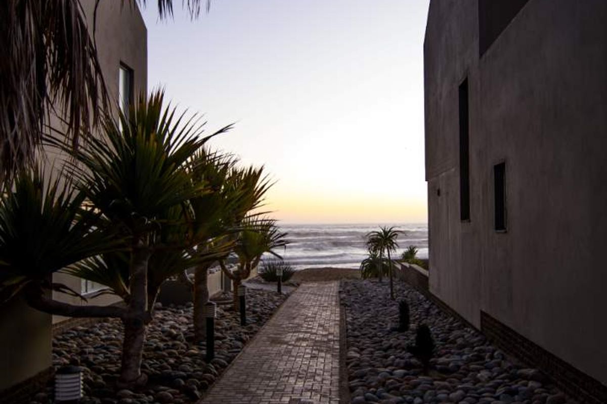 A paved walkway leads between two modern townhouse structures toward a scenic ocean view at sunset.