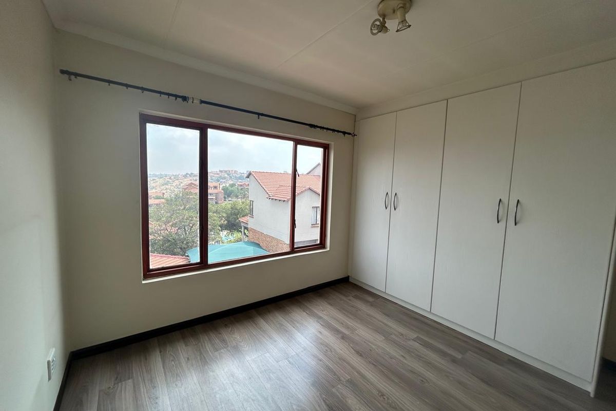 A bright, empty bedroom featuring laminate flooring, built-in cupboards, and a large window offering an elevated view of the surrounding neighborhood.