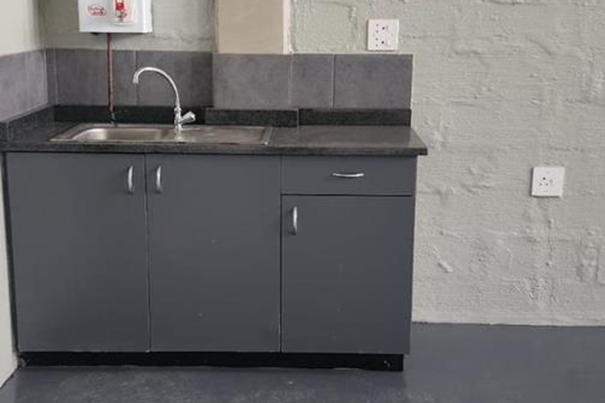 A compact industrial kitchenette featuring a dark grey base cabinet, stainless steel sink, and wall-mounted water heater against a painted concrete block wall.