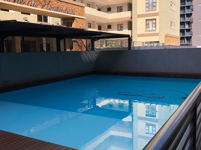 A well-maintained swimming pool area featuring timber decking, a metal balustrade, and a covered patio structure within an apartment complex.