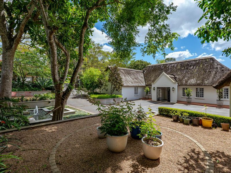 A charming front exterior view featuring a traditional thatched roof, a paved driveway, and a beautifully landscaped garden with a tranquil water feature.